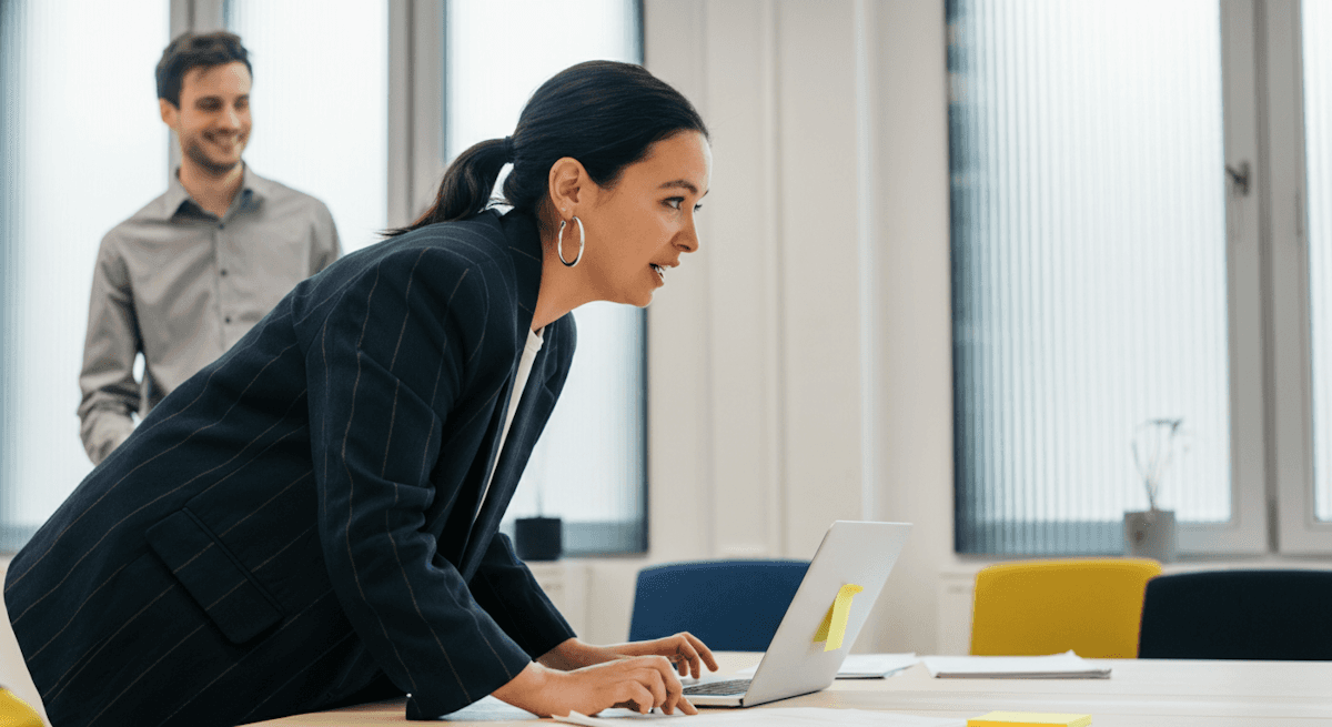A woman leaning over a laptop in a bright office while a colleague looks on